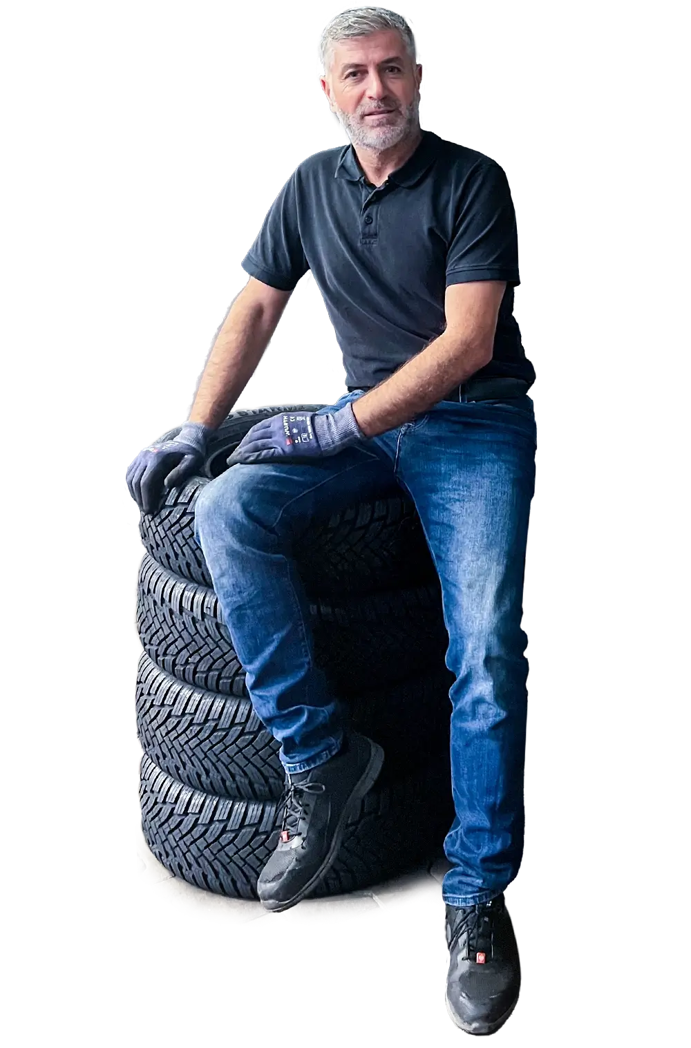 Eine Person in schwarzem Polo-Shirt und blauer Jeans sitzt entspannt auf einem Stapel Reifen, trägt Handschuhe und Sicherheitsschuhe.