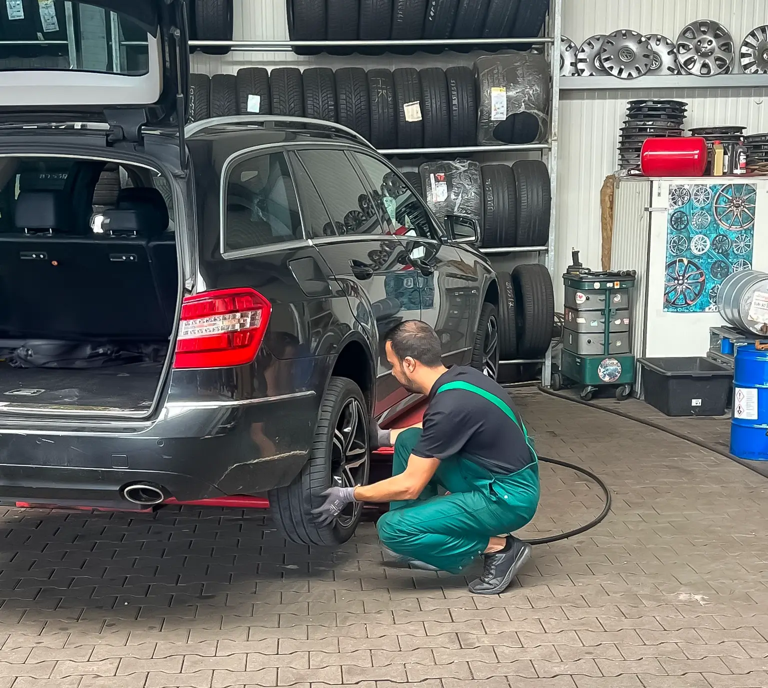 Ein Mechaniker in grüner Arbeitskleidung wechselt oder montiert einen Reifen an einem schwarzen Kombi in einer Werkstatt mit gestapelten Reifen und Felgen im Hintergrund.